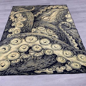 K&ouml;nnte beinhalten: Eine schwarze und goldene Wandteppich mit einer detaillierten Illustration eines Kraken mit ausgebreiteten Tentakeln. Der Krake ist in einem Vintage-Stil mit komplizierten Linien und Mustern dargestellt.