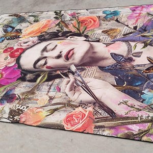 May include: A rectangular art piece featuring a portrait of a woman surrounded by colorful flowers and butterflies. The woman is holding paintbrushes. The artwork has a collage-like design with newspaper clippings in the background.