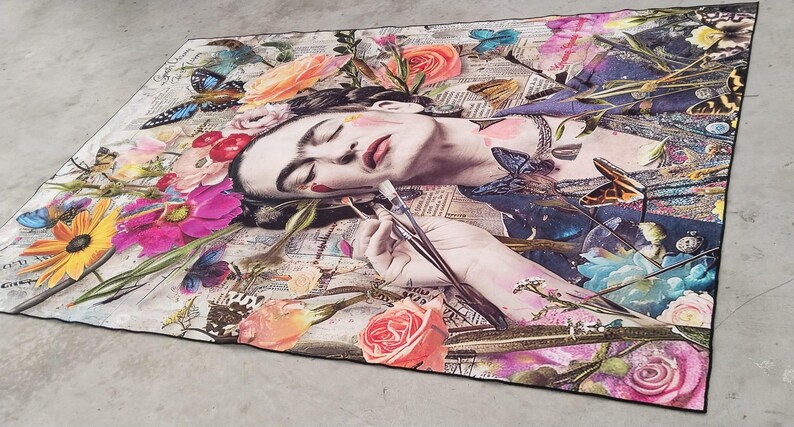 May include: A rectangular art piece featuring a portrait of a woman surrounded by colorful flowers and butterflies. The woman is holding paintbrushes. The artwork has a collage-like design with newspaper clippings in the background.
