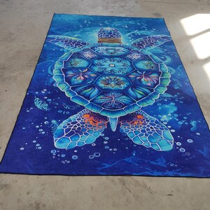Op de afbeelding: Een blauwe en groene mandala-gepatroneerde zeeschildpad op een blauwe achtergrond met witte bubbels. De schildpad is omringd door een galactisch patroon.