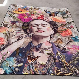 May include: A colorful rug featuring a portrait of a woman with flowers in her hair, butterflies, and a paintbrush. The rug has a black border and is placed on a concrete surface. The artwork is a collage of images and colors.