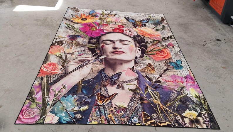 May include: A colorful rug featuring a portrait of a woman with flowers in her hair, butterflies, and a paintbrush. The rug has a black border and is placed on a concrete surface. The artwork is a collage of images and colors.