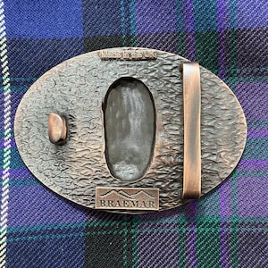 May include: A bronze belt buckle with a textured surface and a raised inscription that reads "Braemar". The buckle also has a smaller inscription that reads "Made in the UK".