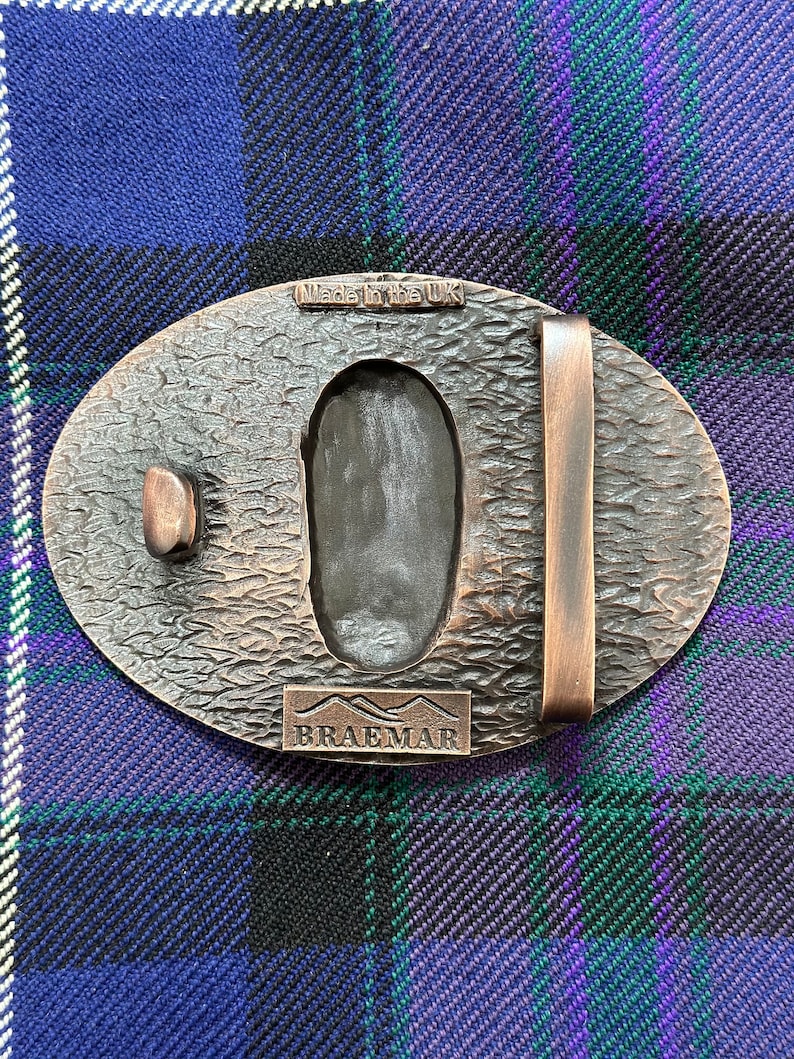 May include: A bronze belt buckle with a textured surface and a raised inscription that reads "Braemar". The buckle also has a smaller inscription that reads "Made in the UK".