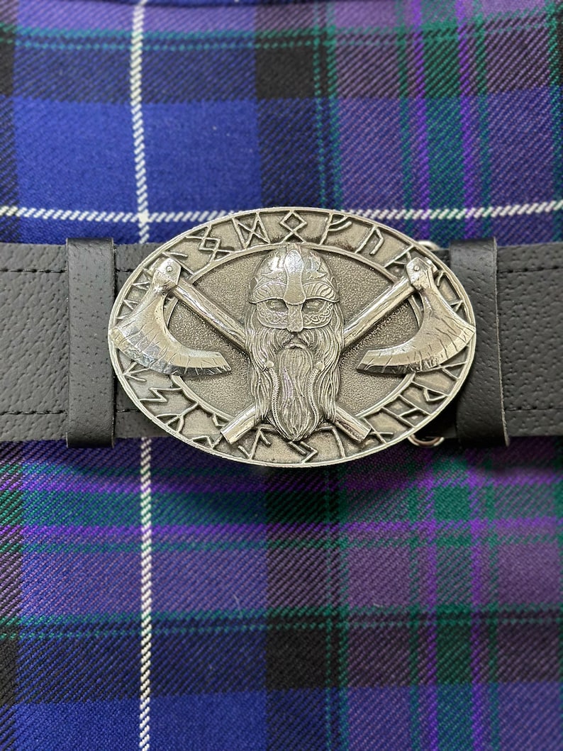 May include: A silver belt buckle with a Viking warrior design. The buckle features a bearded warrior with a helmet and crossed axes. The buckle is surrounded by a circle of runes.