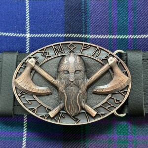 May include: A bronze belt buckle with a Viking warrior design. The buckle features a bearded warrior wearing a horned helmet, holding two crossed axes. The buckle is surrounded by a circle of runes.