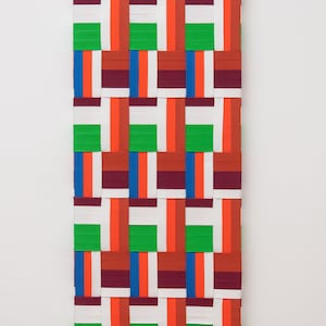 Op de afbeelding: Een geweven wandkleed met een geometrisch patroon van oranje, groene, blauwe, witte en bruine strepen.