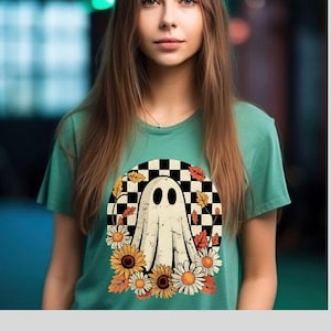 Puede incluir: Una camiseta verde azulado con un diseño de fantasma caprichoso. El fantasma está situado sobre un fondo a cuadros en blanco y negro, rodeado de girasoles, margaritas y hojas de otoño. El diseño tiene una estética vintage con temática otoñal.