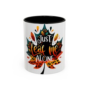 May include: White ceramic mug with a black interior, featuring a colorful leaf design. The leaf is decorated with orange, yellow, and teal accents, and the text "Just leaf me alone" is written on the leaf.