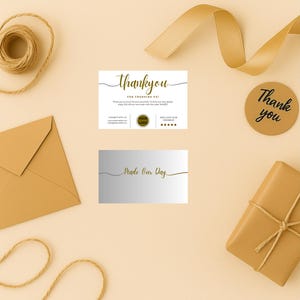 Puede incluir: Una composición plana con elementos de papelería y embalaje en tonos dorados y beige. Incluye una tarjeta de agradecimiento, una caja de regalo atada con hilo, un sobre, una pegatina redonda y un carrete de hilo. La tarjeta dice "Thank you".