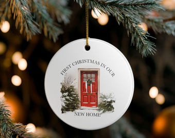 May include: A white ceramic Christmas ornament with the text "FIRST CHRISTMAS IN OUR NEW HOME." The ornament features a red front door with a wreath, snow, and evergreen trees.