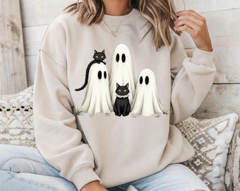 Puede incluir: Sudadera beige con un diseño de Halloween. Presenta tres fantasmas blancos y dos gatos negros. Los gatos están sobre la hierba y los fantasmas tienen ojos negros. Un diseño divertido y festivo para la temporada de miedo.