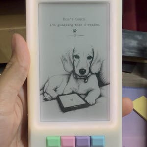 Könnte beinhalten: Eine weiße E-Reader-Hülle mit leuchtendem Rand und vier pastellfarbenen Tasten in Grün, Rosa, Lila und Blau. Der Bildschirm zeigt eine schwarz-weiße Skizze eines Welpen mit dem Text: Don't touch. I'm guarding this e-reader.