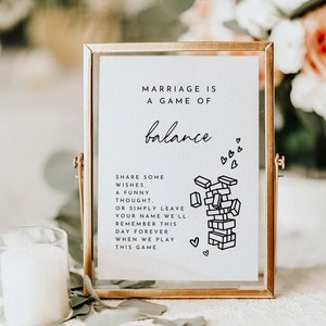 Puede incluir: Un cartel enmarcado en dorado con el texto "El matrimonio es un juego de equilibrio" y una ilustración en blanco y negro de un juego de Jenga. El cartel también incluye el texto "Comparte algunos deseos, un pensamiento divertido, o simplemente deja tu nombre. Recordaremos este día para siempre cuando juguemos a este juego."