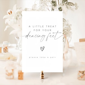 May include: A white sign with black text that reads "A little treat for your dancing feet" with a small heart symbol below the text and "please take a pair" below the heart. The sign is on a gold stand and there are white flowers in the background.