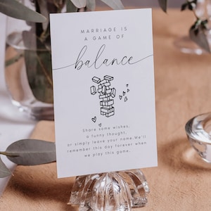 May include: White card with black text that reads "Marriage is a game of balance" with a black and white illustration of a tower of blocks falling over. The card is sitting on a clear glass stand.