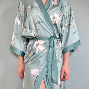 Könnte beinhalten: Eine tealblaue Satin-Robe mit einem Blumendruck aus weißen Magnolienblüten. Die Robe hat einen Selbstgürtel und eine lange, fließende Silhouette.