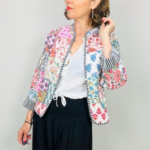 Pu&ograve; includere: Una giacca bianca con motivo floreale e bordini a righe bianche e nere. La giacca &egrave; realizzata con un patchwork di tessuti diversi e ha un colletto alla coreana. La modella indossa la giacca sopra una maglia bianca e pantaloni neri.