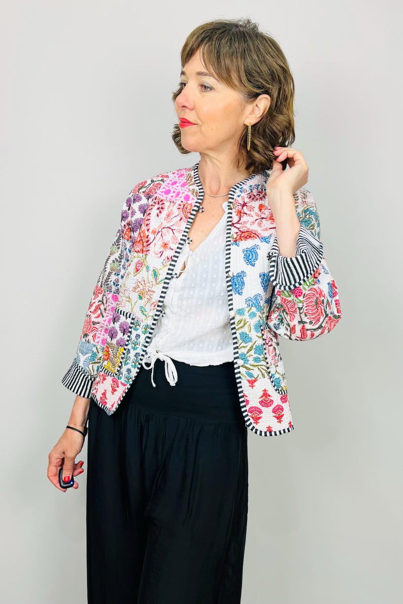 Pu&ograve; includere: Una giacca bianca con motivo floreale e bordini a righe bianche e nere. La giacca &egrave; realizzata con un patchwork di tessuti diversi e ha un colletto alla coreana. La modella indossa la giacca sopra una maglia bianca e pantaloni neri.