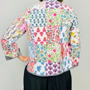Pu&ograve; includere: Una giacca trapuntata bianca con un design a patchwork che presenta motivi floreali in vari colori, tra cui rosa, blu, verde e viola. La giacca ha un colletto alla coreana e maniche lunghe.