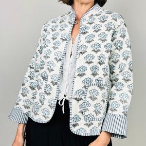 Op de afbeelding: Een witte gewatteerde jas met een blauw bloemenpatroon. De jas heeft een opstaande kraag, driekwartmouwen en twee voorzakken. De manchetten en de bies hebben blauw-witte strepen. De jas is aan de voorkant open.