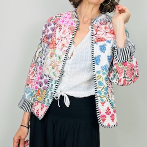 Pu&ograve; includere: Una giacca trapuntata bianca con un design a patchwork che presenta motivi floreali colorati. La giacca ha un colletto alla coreana e un bordo a righe bianco e nero sulle maniche.
