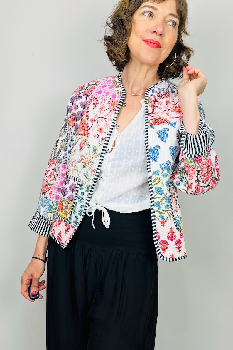 Pu&ograve; includere: Una giacca trapuntata bianca con un design a patchwork che presenta motivi floreali colorati. La giacca ha un colletto alla coreana e un bordo a righe bianco e nero sulle maniche.