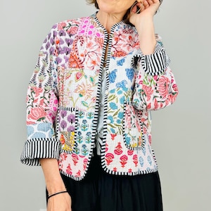 Pu&ograve; includere: Una giacca trapuntata bianca con un design patchwork floreale colorato. La giacca ha un colletto alla coreana, maniche lunghe e un bordo a righe bianco e nero.