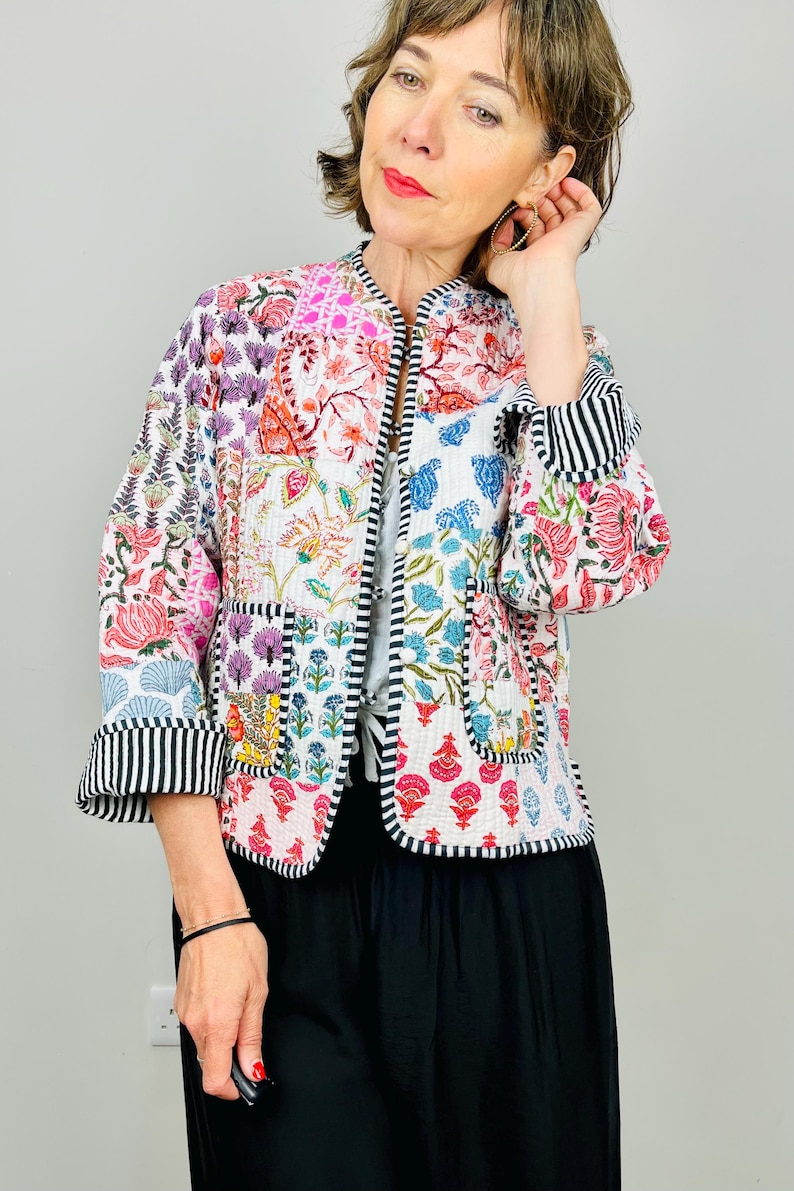 Pu&ograve; includere: Una giacca trapuntata bianca con un design patchwork floreale colorato. La giacca ha un colletto alla coreana, maniche lunghe e un bordo a righe bianco e nero.