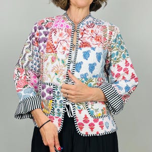 Pu&ograve; includere: Una giacca trapuntata bianca con un design a patchwork che presenta motivi floreali e paisley in tonalit&agrave; di rosa, blu, verde e rosso. La giacca ha un colletto alla coreana, una chiusura a cravatta e bordi a righe bianche e nere.