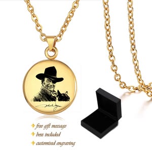 May include: A gold-colored necklace with a round pendant featuring a black and white portrait of a man in a hat and a dog. The necklace includes a matching gold chain and a black gift box.