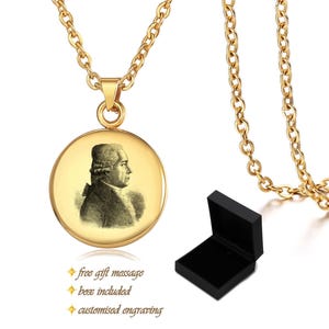 May include: A gold-coloured necklace with a circular pendant featuring a black and white portrait. The necklace includes a matching chain and a black gift box. The pendant has a portrait of a man.