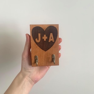 以下が含まれることがあります： 中央に「J+A」の文字が刻印されたダークブラウンのハート型インレイが特徴の長方形の木製ウォールフック。明るい色調の木製ベースの下部には、物を掛けるための小さな真鍮色の金属製フックが2つ取り付けられています。