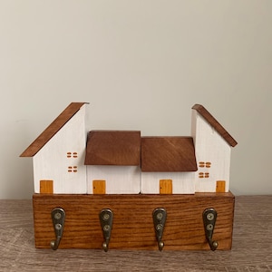 May include: A wooden wall-mounted key holder featuring four metal hooks attached to a rectangular base. Above the base are four miniature house structures with white walls, brown roofs, and small painted orange doors and windows.