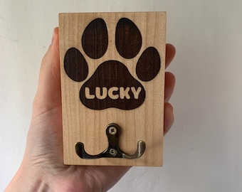 May include: Wooden wall hook featuring a dark brown paw print and the word "LUCKY". It has a double bronze hook for hanging items. The wood has a light, natural finish. The hook is designed for wall mounting.