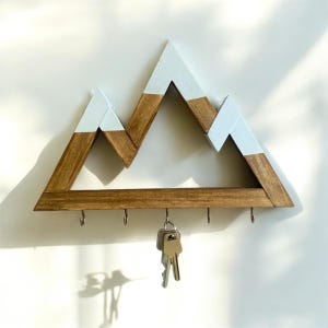 May include: A wall-mounted wooden key holder shaped like a mountain range with three peaks. The lower sections are stained a natural brown wood tone, while the upper tips are painted white. Five metal hooks are attached to the base, with one set of keys.
