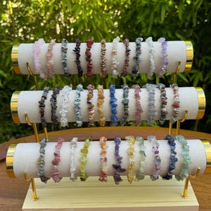 Peut inclure: Présentation de bracelets de pierres précieuses de différentes couleurs, notamment rose, marron, vert et violet. Les bracelets sont disposés sur un présentoir à trois niveaux doré et crème, sur fond vert naturel.