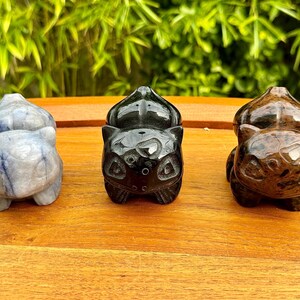 May include: Three carved stone figurines in the shape of a Bulbasaur. The figurines are in shades of blue, black, and brown. The figurines are on a wooden surface with green foliage in the background.