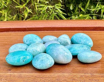 May include: A collection of smooth, oval-shaped stones in varying shades of turquoise and blue. The stones are arranged on a wooden surface, with green foliage visible in the background. The stones appear to be polished and have a natural, organic look.