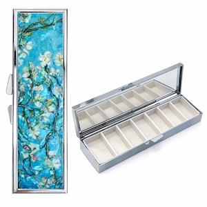 以下が含まれることがあります： ヒンジ付きの蓋と鏡面仕上げのインテリアを備えたシルバーのピルケース。蓋には、青い背景に白い花が描かれた絵画が施されています。内部には、クリーム色の生地で裏打ちされた複数のコンパートメントがあります。