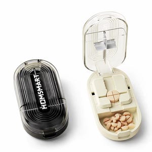 Op de afbeelding: Twee pillenorganizers. Eén is gesloten, zwart met de tekst "HOMSMART". De andere is open, gebroken wit, met een pillensnijder en verschillende gesneden pillen. De deksel is transparant.