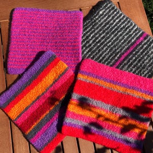 Könnte beinhalten: Vier farbenfrohe, quadratische Filzartikel aus Wolle sind auf einer Holzoberfläche angeordnet. Die Artikel weisen leuchtende Farben auf, darunter Fuchsia, Rot, Orange, Lila und Grau. Ein Artikel hat horizontale Streifen, während andere einfarbige oder gestreifte Muster aufweisen.
