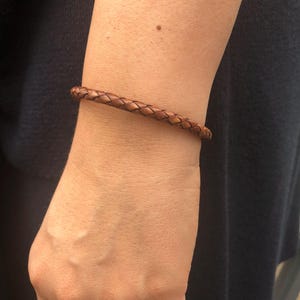 Könnte beinhalten: Ein braunes, geflochtenes Lederarmband. Das Armband wird am Handgelenk getragen. Das Leder hat eine mittelbraune Farbe und scheint aus mehreren miteinander verflochtenen Strängen zu bestehen. Das Armband ist ein schlichtes, klassisches Design.