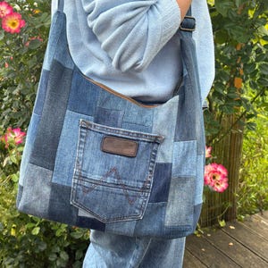 Könnte beinhalten: Eine Patchwork-Jeanstasche aus verschiedenen blauen Denim-Stoffquadraten. Die Tasche verfügt über eine Außentasche mit einem braunen Lederaufnäher der Marke Wrangler und charakteristischen W-Nähten sowie einen verstellbaren Schulterriemen.