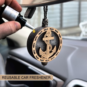 May include: A reusable car air freshener with a wooden anchor design. The round, dark-colored freshener has a light-colored wooden frame with an anchor cutout. A hand is applying a liquid from a small bottle to the freshener. The text "REUSABLE CAR FRESHENER" is at the bottom.