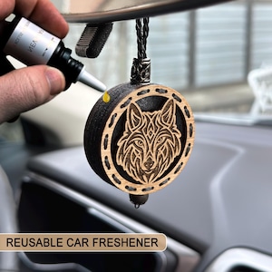 Puede incluir: Un ambientador de coche redondo de madera con un diseño de lobo cortado con láser. El ambientador es negro con una imagen de lobo marrón claro y un cordón negro trenzado. El texto "REUSABLE CAR FRESHENER" está en la parte inferior.