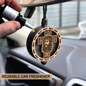May include: A reusable car freshener with a wooden cross design featuring an image of Jesus. The round, dark-coloured freshener has a light-coloured wooden cross with floral accents and a black cord for hanging. The text "REUSABLE CAR FRESHENER" is visible.