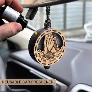 Puede incluir: Un ambientador de coche redondo de madera, con un borde negro y un anillo exterior de madera clara. El diseño presenta manos rezando y el texto "Drive Safe - God is With You". Se adjunta un cordón negro para colgar.