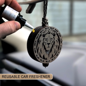 May include: A circular, dark wooden car air freshener featuring a lion's head design. It hangs from a black braided cord. The words "REUSABLE CAR FRESHENER" are displayed on a beige banner at the bottom of the image.