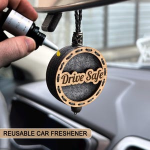 May include: A circular car air freshener with the words "Drive Safe" cut into the wood. The freshener is made of wood and grey felt. A hand is shown applying a liquid from a small bottle to the freshener. The words "REUSABLE CAR FRESHENER" are at the bottom.